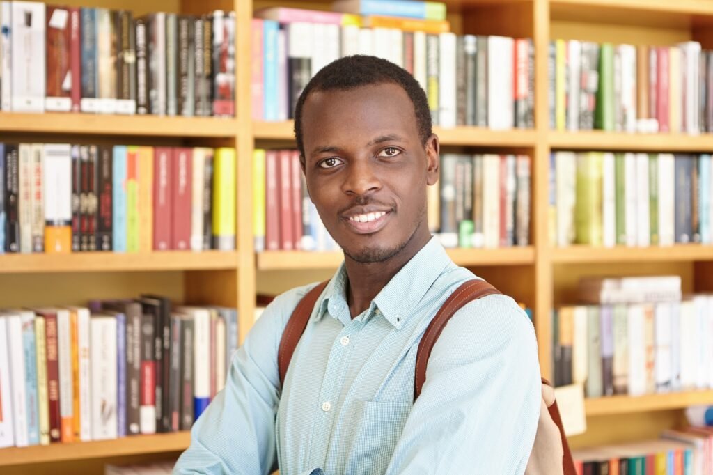 Black college student wearing elegant shirt holding rucksack isolated over library background lookin